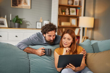 A couple sits on a comfortable sofa in their cozy living room, sharing a moment as they look at a...