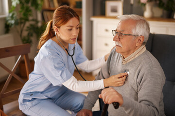 Fototapeta premium During a home visit, a nurse is examining a senior man with a stethoscope to check his heartbeat. The man is sitting comfortably in his living room with a cane.