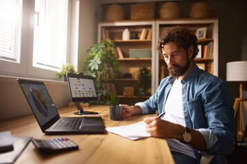 Fototapeta premium A man sits at a wooden desk in a well-lit home office, engaged in writing notes while drinking from a cup. He is surrounded by a laptop, a tablet, and house plants, indicating a productive workspace.