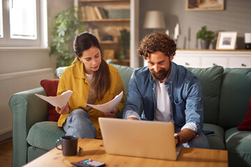 A couple sits together on a couch in their living room, with one partner reviewing life insurance documents while the other works on a laptop. The atmosphere is relaxed and productive.
