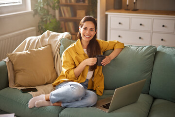 Woman in a yellow jacket relaxes on a sofa with a coffee cup, smiling at her laptop in a bright and...