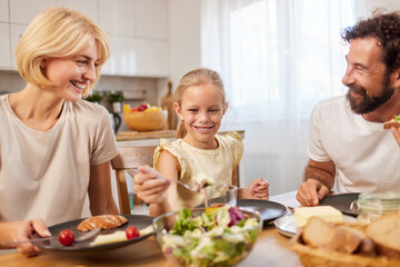 Three family members gather around a table filled with food, happily sharing a meal. A child beams with joy as the adults engage in conversation and enjoy their dishes.