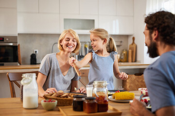 Family members gather around a wooden table in a bright kitchen, laughing and sharing breakfast...