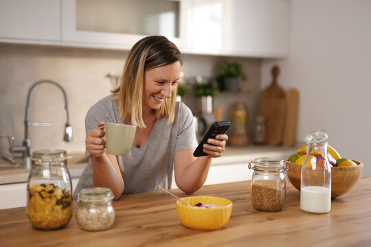 A woman sits at a wooden kitchen table, smiling as she looks at her smartphone. She is holding a cup of coffee and has a bowl of cereal in front of her, with jars of food nearby.