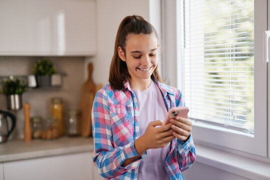 A teenage girl with brown hair is smiling as she checks her smartphone. She is in a cozy kitchen with sunlight streaming through the window. - Powered by Adobe