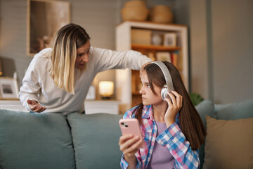 A teenage girl wears headphones and looks at her phone, seemingly uninterested, while her mother...