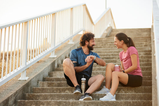 Two individuals take a break from their outdoor exercise routine, sitting on stairs while sharing a light moment and enjoying refreshing drinks under the warm sun.