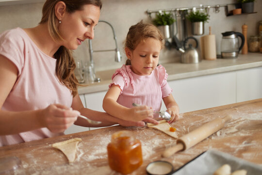 A mother and her young daughter are happily engaged in baking together at a kitchen counter, working with dough and jam in a bright, inviting space.