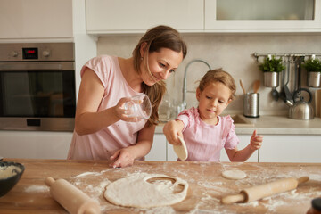 In a bright and inviting kitchen, a mother guides her young daughter as they roll dough, fostering...