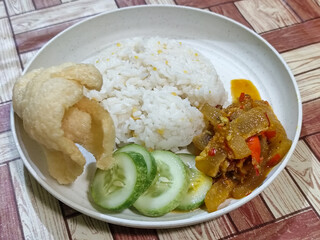 A serving of mixed rice, warm white rice accompanied by stir-fried spicy kikil with sliced red chilies and fresh cucumber on a white plate, served on a checkered wooden table.