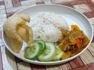 A serving of mixed rice, warm white rice accompanied by stir-fried spicy kikil with sliced red chilies and fresh cucumber on a white plate, served on a checkered wooden table.