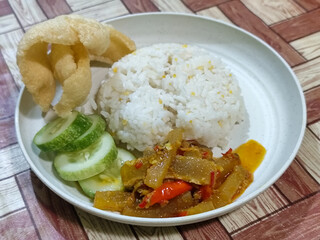 A serving of mixed rice, warm white rice accompanied by stir-fried spicy kikil with sliced red chilies and fresh cucumber on a white plate, served on a checkered wooden table.