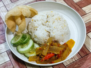 A serving of mixed rice, warm white rice accompanied by stir-fried spicy kikil with sliced red chilies and fresh cucumber on a white plate, served on a checkered wooden table.