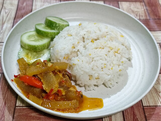 A serving of mixed rice, warm white rice accompanied by stir-fried spicy kikil with sliced red chilies and fresh cucumber on a white plate, served on a checkered wooden table.