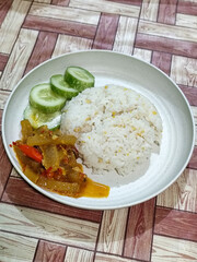 A serving of mixed rice, warm white rice accompanied by stir-fried spicy kikil with sliced red chilies and fresh cucumber on a white plate, served on a checkered wooden table.