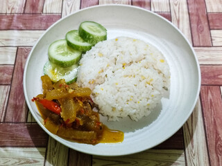 A serving of mixed rice, warm white rice accompanied by stir-fried spicy kikil with sliced red chilies and fresh cucumber on a white plate, served on a checkered wooden table.
