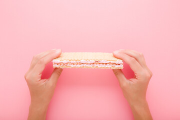 Young adult woman hands holding and showing wafer with white pink cream on light pink table background. Pastel color. Sweet snack. Closeup. Point of view shot. Top down view.