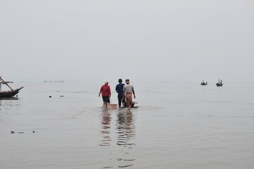 Scene of individuals walking in shallow waters with fishing boats in the distance