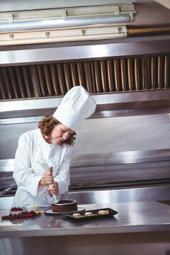 Naklejki Mature pastry chef piping white frosting onto chocolate cake at stainless workbench with cherries