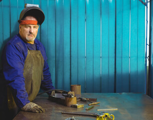 Welder wearing leather apron and helmet operating gas torch near cylinders in turquoise workshop