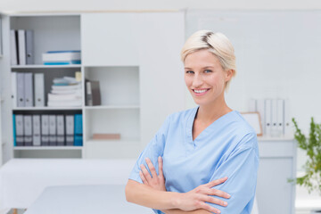 Female nurse wearing blue scrubs standing in exam room featuring shelving unit and exam table