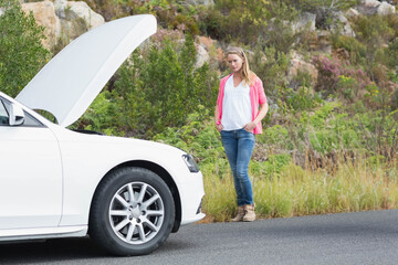 Woman standing on rural roadside beside white sedan with hood open, examining engine compartment