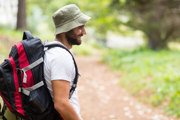 Adult male hiker standing on dirt trail with large red black backpack, bucket hat, copy space