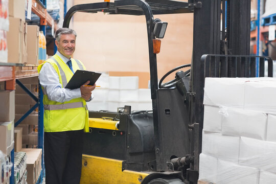Senior man in safety vest inspecting shelves with clipboard next to forklift in warehouse