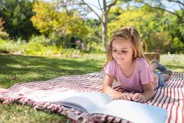 Female child lying on picnic blanket reading open book near basket in park, copy space