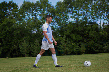 Male soccer player walking across grass field dribbling soccer ball wearing blue jersey and cleats