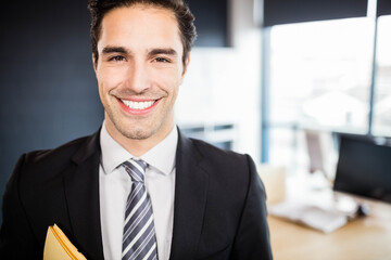 Man wearing suit holding yellow folder smiling at office desk with monitor and papers near windows
