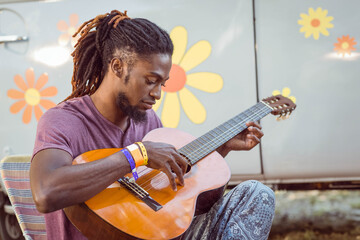 African American man strumming acoustic guitar on striped chair outdoors beside flowered van