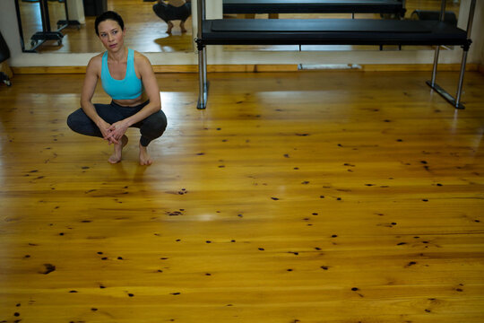 Fototapeta Woman is squatting barefoot on fitness studio floor with reformer bench and mirror reflection