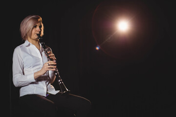 Female musician sitting on stool at stage edge playing clarinet under spotlight, copy space
