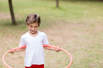 Boy standing in grassy park holding pink plastic hula hoop behind back on sunny day