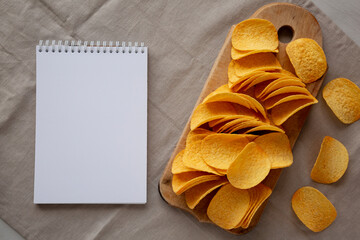Bacon flavoured potato chips on a wooden board, blank notepad, top view.