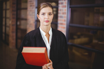 Woman in black robe holding large red book in office corridor with brick columns