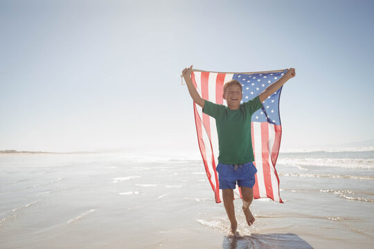 Boy running through shallow ocean water on beach holding American flag billowing under clear sky