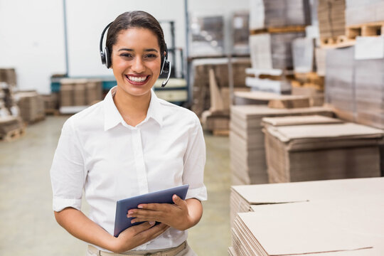 Woman wearing headset and shirt standing in warehouse holding tablet monitoring forklift and boxes