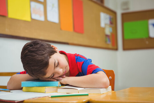 School-age boy sleeping on textbook stack at classroom desk with pencil and open workbook