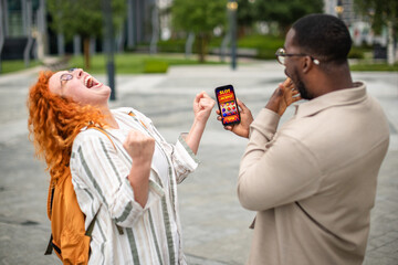 Excited woman celebrates winning as her friend watches on smartphone in urban park setting