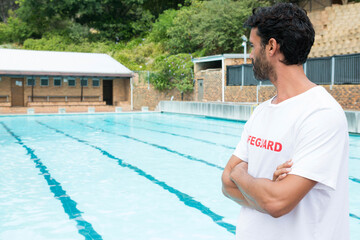 Lifeguard standing watch at edge of swimming pool, with lane markings and brick changing rooms