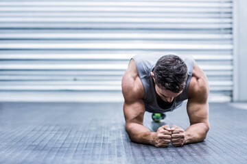 Adult male athlete holding forearm plank position on textured gym floor in front of metal shutter