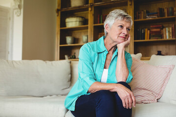 Senior woman sitting on sofa wearing turquoise shirt, gazing to right at home with wooden bookshelf