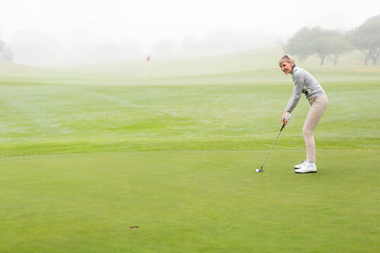 Senior woman lining up putt by flag on dew-covered green with putter and ball, copy space - Powered by Adobe