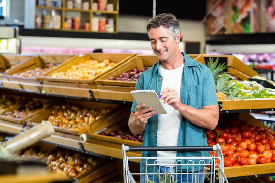 Middle-aged man writing notes on spiral-bound notepad in supermarket produce aisle by shopping cart - Powered by Adobe