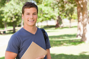 Male student wearing blue T-shirt on campus lawn, holding folder and backpack under sunlight
