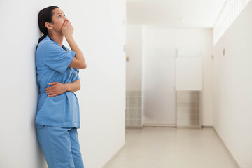 Female healthcare worker leaning in hospital corridor wearing scrubs with stethoscope, copy space