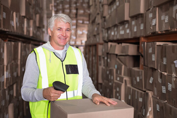 Male warehouse worker scanning box with barcode scanner in narrow aisle wearing neon safety vest
