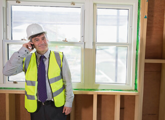 Senior man inspecting window frame inside building with hard hat safety vest and smartphone